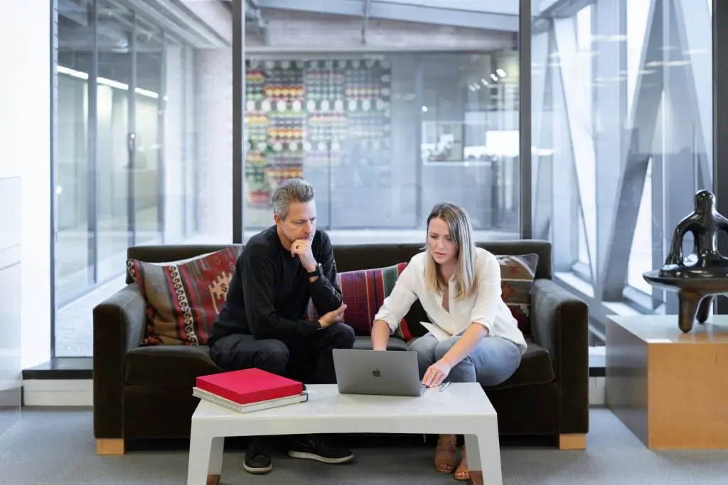 a man and a woman sit on a couch looking at a laptop